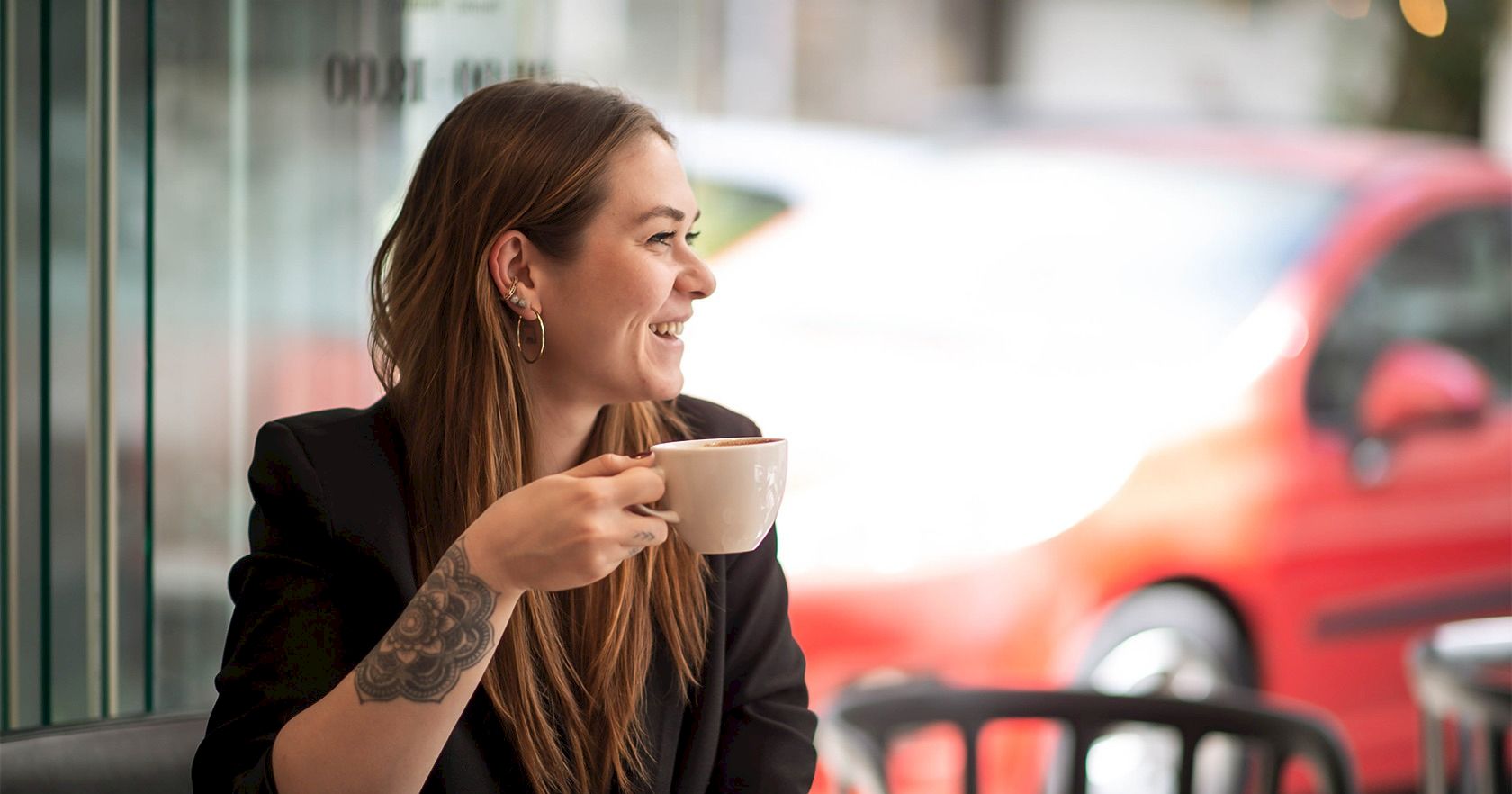 A smiling woman with long hair sits at a cafe table, holding a cup of coffee; a plate with pastries is in front, outdoors on a sunny day.