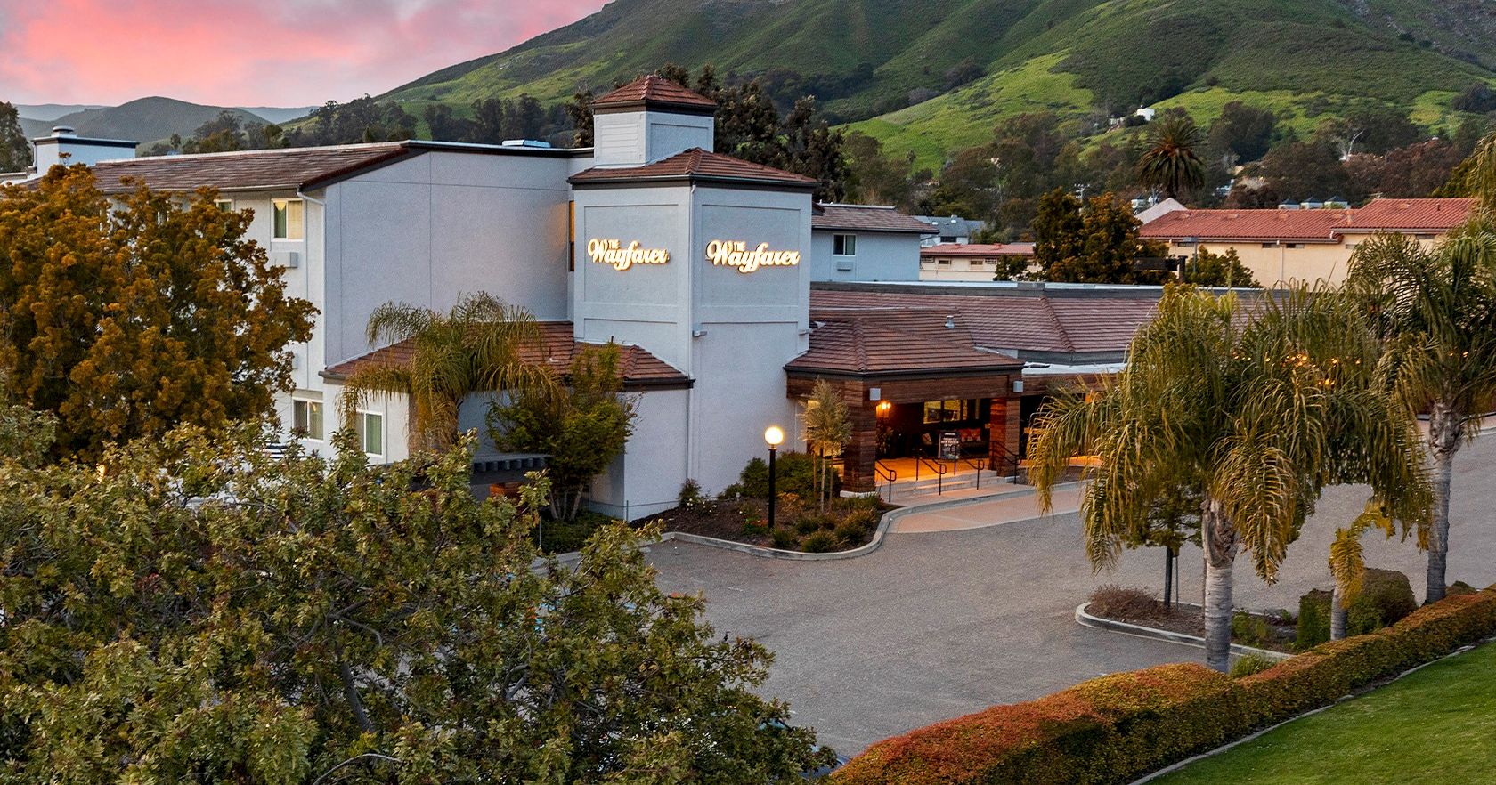A view of a hotel with a light blue facade, palm trees, and a signage reading &ldquo;Hilton&rdquo; or similar, set against green hills at sunset.
