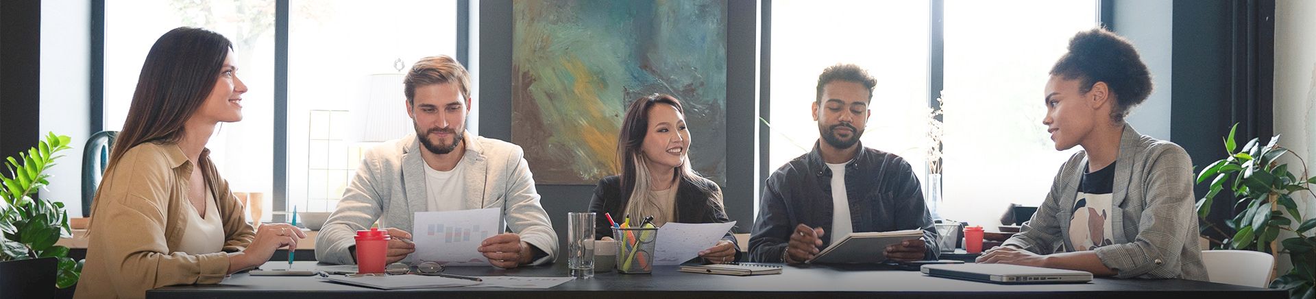 A diverse group of five adults sit around a long table in a bright office, collaborating with papers, laptops, and coffee cups.