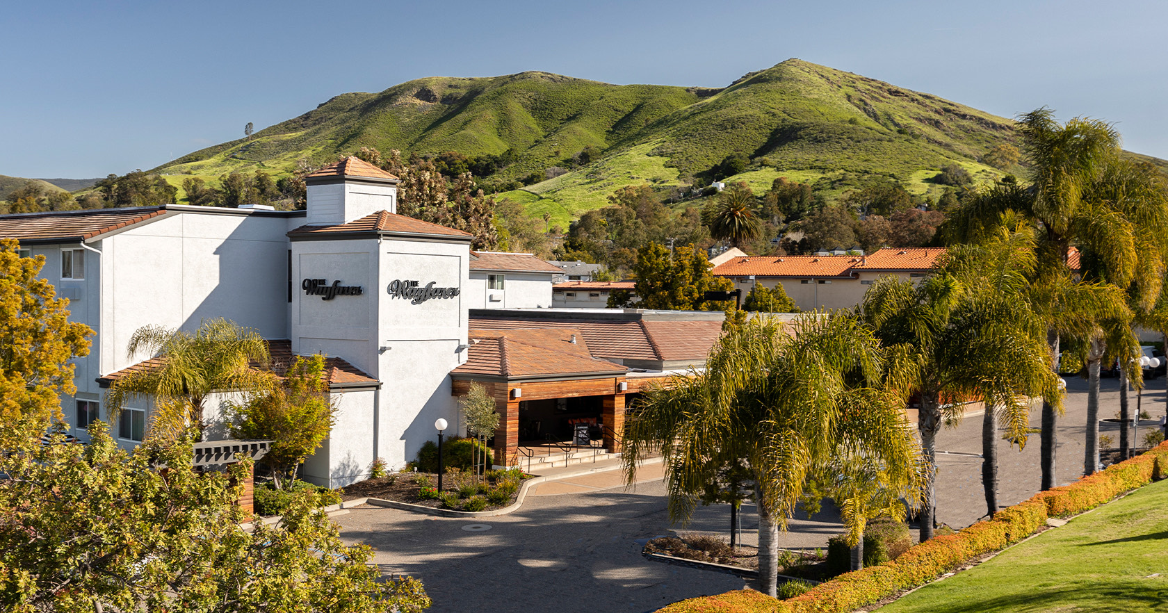 A resort or hotel with white buildings, palm trees, and a gated entrance set against green hills.