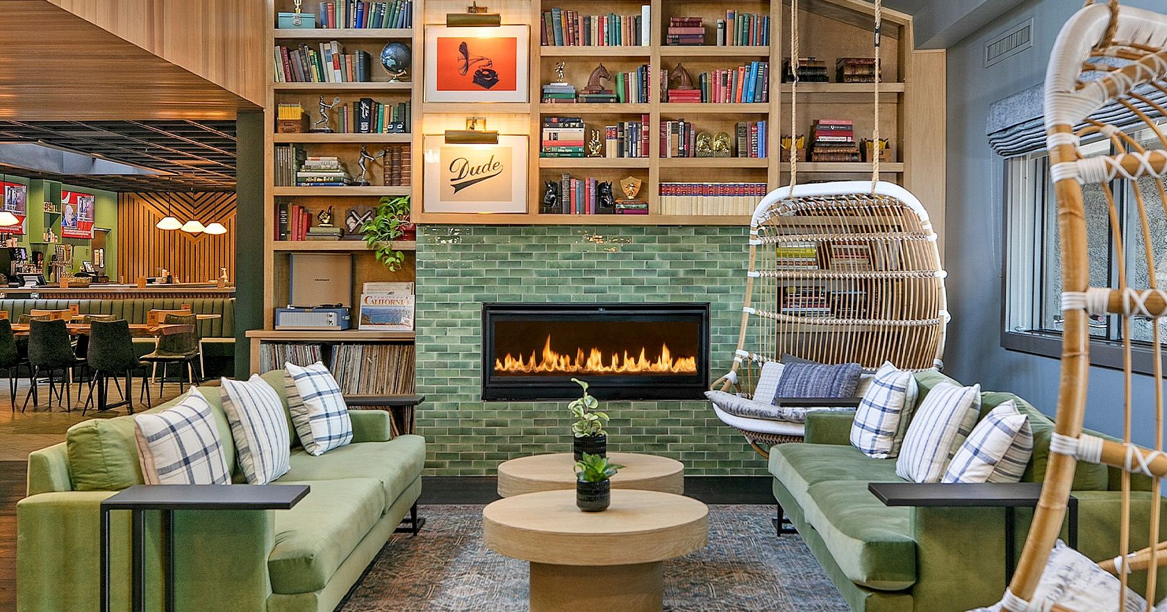 A cozy, modern lounge with green sofas, a brick fireplace, floor-to-ceiling bookshelves, and a hanging wicker chair&mdash;perfect for reading.