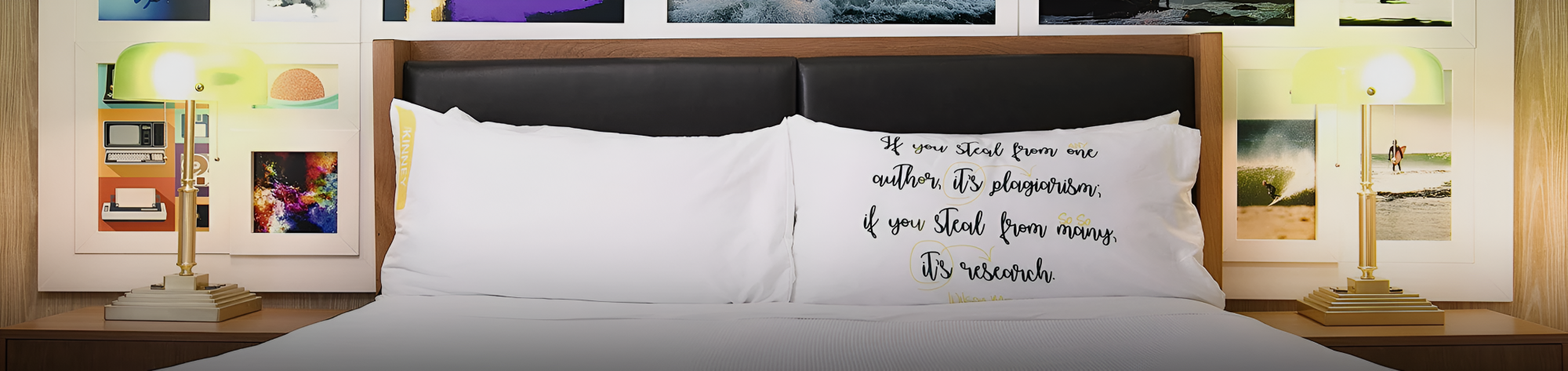 A neatly made bed with white pillows, black headboard, and art-filled wall. Two lamps glow on either side, a cozy, gallery-like bedroom.