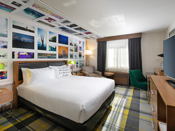 A bright hotel room with a large bed, colorful collage wall, two nightstands, a sitting area, a desk with TV, and plaid carpet. Ends here.