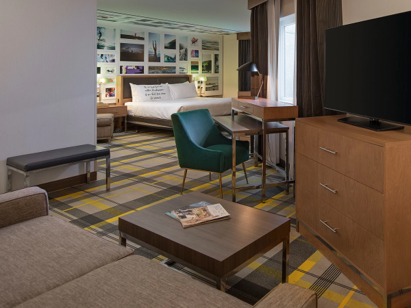 A modern hotel suite with a bed, seating area, desk, a large TV on a wooden dresser, colorful plaid carpet, and framed photos on the wall.