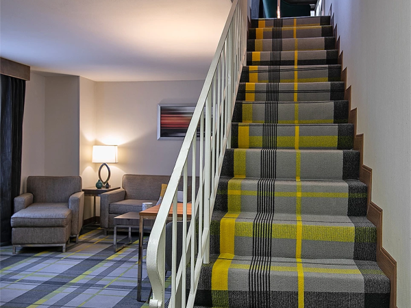 A modern hotel staircase with a colorful striped carpet leads upstairs, adjacent seating with chairs and a lamp in a cozy lounge area.