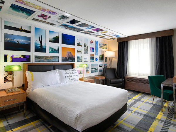 A stylish hotel room with a gallery wall of photos above the bed, a bright white bed, nightstands, a seating area, and a plaid yellow-gray carpet.