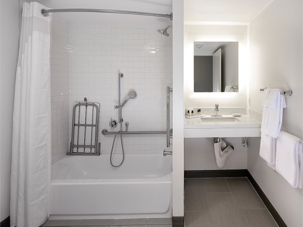 A modern bathroom with a white tile shower/bathtub, a handheld shower, a towel rack, and a floating vanity with a backlit mirror.