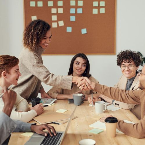 A diverse team around a table high-fives and smiles after a meeting; sticky notes on a board, laptops, coffee cups, teamwork vibe.
