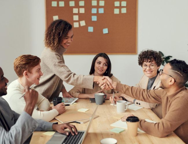 A diverse team around a table high-fives and smiles after a meeting; sticky notes on a board, laptops, coffee cups, teamwork vibe.