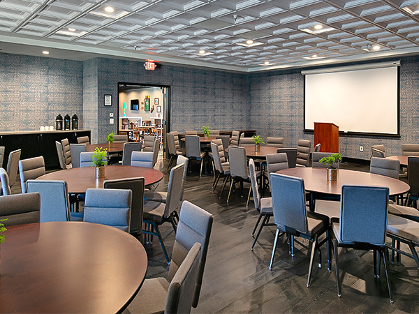 A conference or banquet room with round and rectangular tables, blue-gray chairs, small potted plants, a projector screen, and a doorway to a coffee station; ceiling panels and soft lighting.