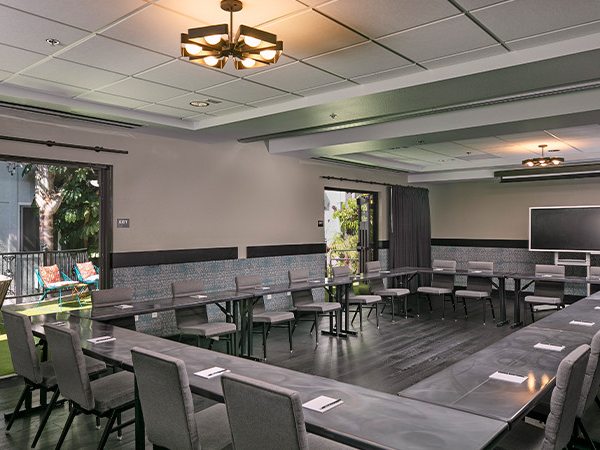 A large conference room with U-shaped and classroom-style seating, gray chairs, long tables, a chalkboard/whiteboard, and outdoor view.