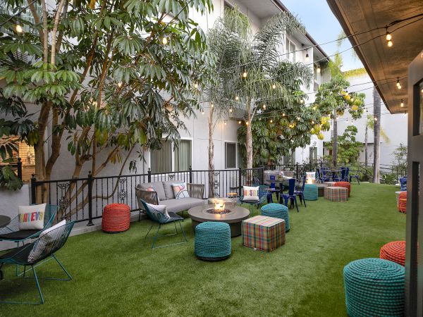 A cozy outdoor lounge with string lights, colorful poufs, low tables, and a grassy courtyard surrounded by trees and modern apartments.