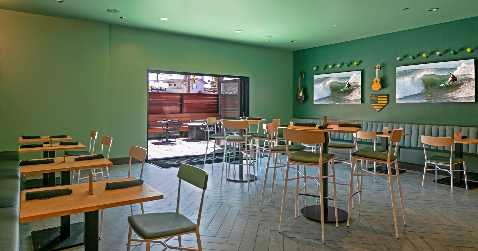 A bright, modern caf&eacute; interior with teal walls, wooden tables and chairs, ceiling lights, a view to an outdoor area, and framed wave art on the wall.