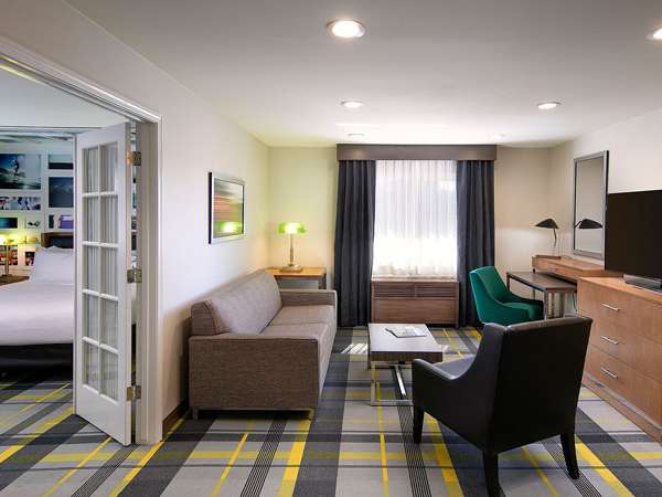 A modern hotel suite with a seating area: sofa, coffee table, two chairs, a desk with a TV, and a bed visible through an open doorway in a bright, stylish room.