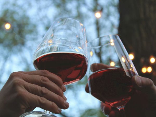 Two people clink wine glasses outdoors, red wine, festive lights in the background, hands up in a toast.
