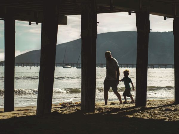 Two people, likely a parent and child, walk along a beach under a wooden pier as the sun sets near the water.