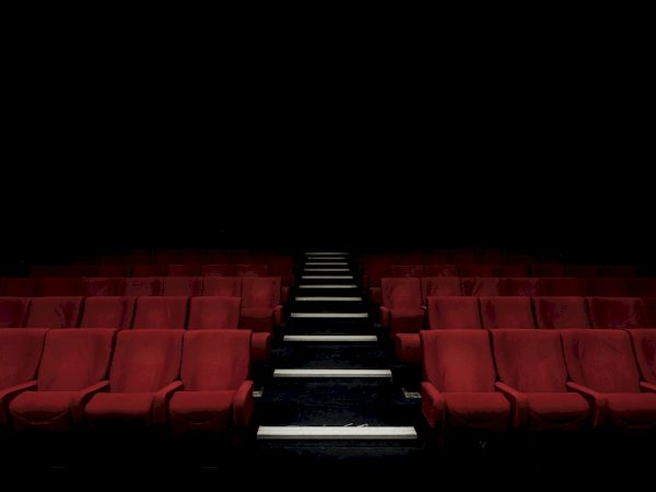 A dim red theater with empty seats and a single illuminated aisle steps leading toward the dark stage, creating a mysterious, silent mood.