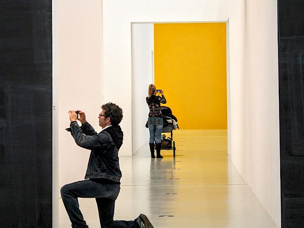 A man kneels and photographs a doorway in a gallery; multiple doorways recede into a yellow room.