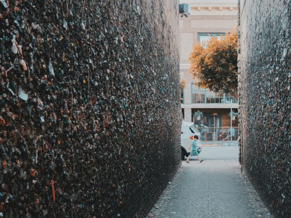 A narrow alley with tall, dark walls on both sides, a wet cobblestone path, and a hint of a car and building in the background, sunny day.