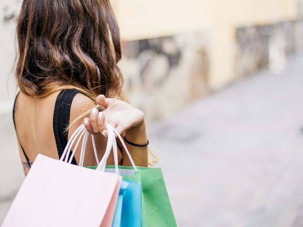 A person walking away with colorful shopping bags in hand, wearing a black top and wavy hair, on a bright sidewalk, shopping spree vibes.