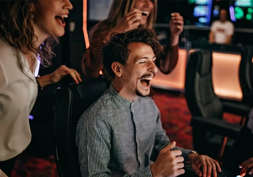 People celebrate at a gaming or casino venue; a man laughs joyfully while others cheer around him, creating a lively, social scene.