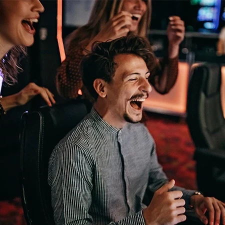 People celebrate at a gaming or casino venue; a man laughs joyfully while others cheer around him, creating a lively, social scene.