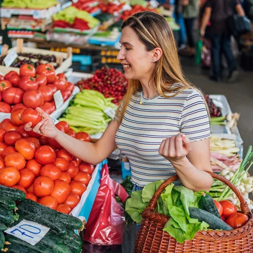 A smiling woman shops for tomatoes and fresh produce at a vibrant outdoor market, holding a basket and selecting veggies. End.