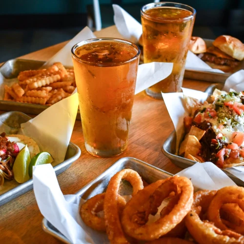 A table of Mexican food with tacos, fries, onion rings, and two iced drinks; vibrant toppings and plates ready to share, delicious.