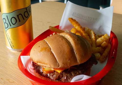 A fast-food meal: a cheeseburger on a red tray with fries, plus a can of Blonde beer beside the tray, all on a wooden table.