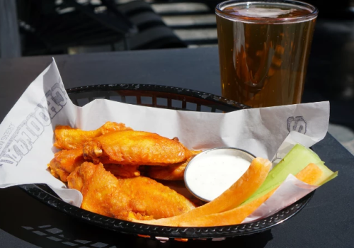 Basket of buffalo wings with dipping sauce, melon slices, and a glass of beer on a dark table.