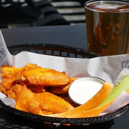 Basket of buffalo wings with dipping sauce, melon slices, and a glass of beer on a dark table.