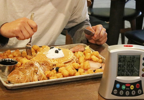 A breakfast feast: waffles with whipped cream, pancakes, golden hash browns, syrup, and a person texting while timer beeps.