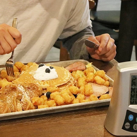 A breakfast feast: waffles with whipped cream, pancakes, golden hash browns, syrup, and a person texting while timer beeps.