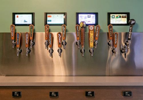 A row of beer taps with digital screens above, lined up on a stainless bar, ready for pours.