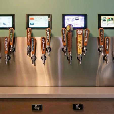 A row of beer taps with digital screens above, lined up on a stainless bar, ready for pours.