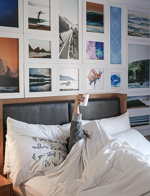 A person in bed under white bedding holds up a phone, with a wall of framed photos and art behind, creating a cozy, photo-filled bedroom vibe.