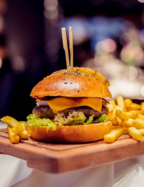 A juicy burger with cheddar, lettuce, onion, and sesame bun on a wooden board, served with fries, skewered with two sticks, delicious.