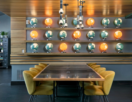 A modern dining space with a long wooden table, yellow chairs, and circular wall shelves displaying orange-lit globes against a striped wood backdrop.