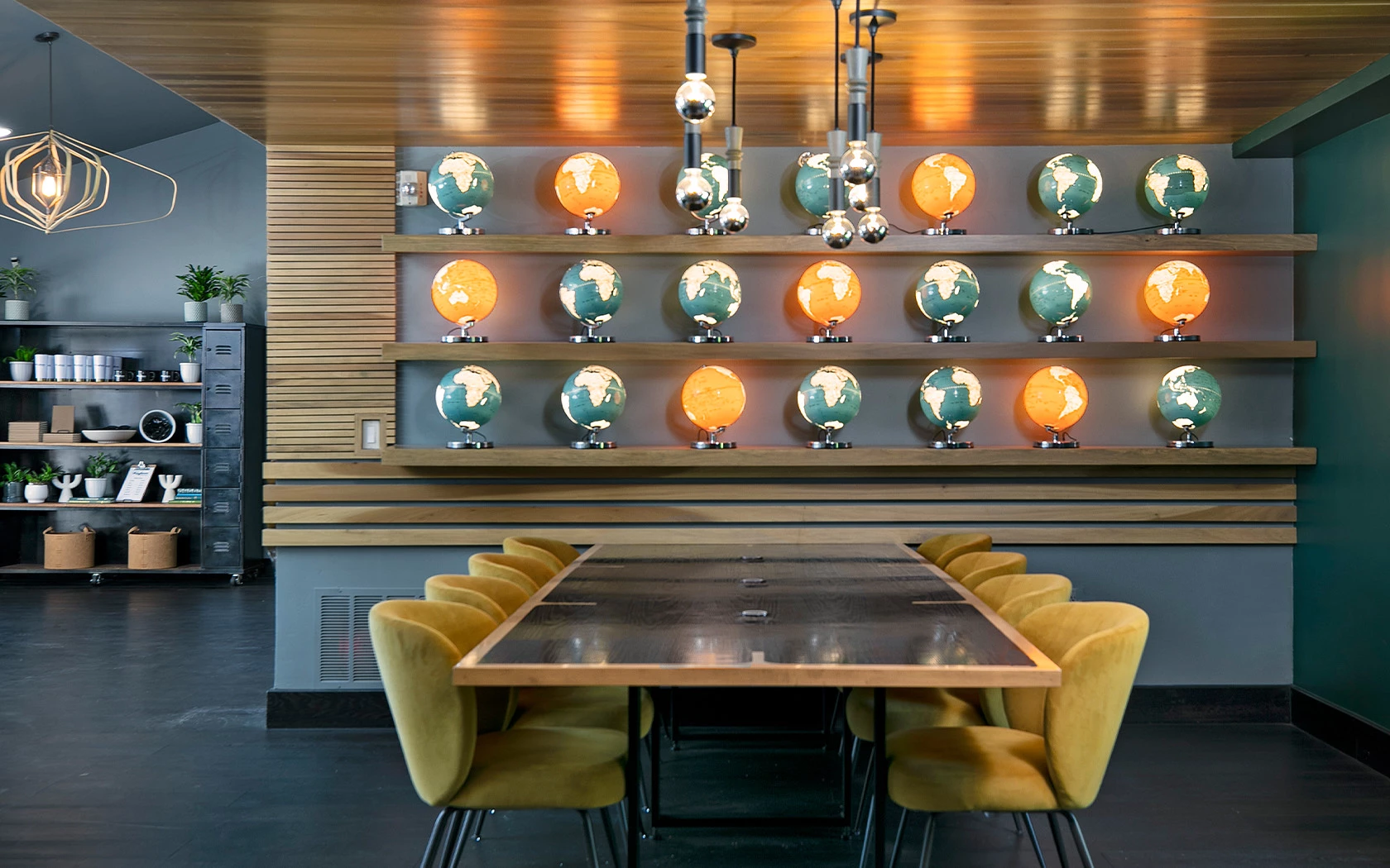 A modern dining space with a long wooden table, yellow chairs, and circular wall shelves displaying orange-lit globes against a striped wood backdrop.