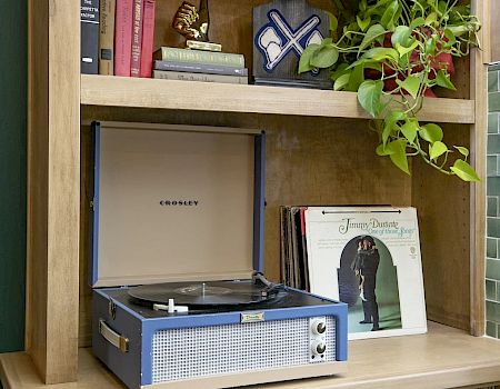 A wooden desk with a record player on the lower shelf, books and a potted plant above, and vinyl records stored beneath the shelf.