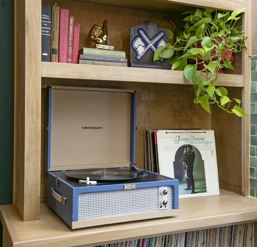 A wooden desk with a record player on the lower shelf, books and a potted plant above, and vinyl records stored beneath the shelf.