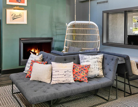 Cozy modern living room with a tufted gray sofa, colorful patterned pillows, a hanging chair, fireplace, and bookshelves by a window.
