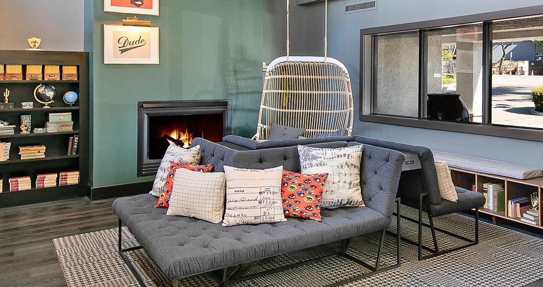 Cozy modern living room with a tufted gray sofa, colorful patterned pillows, a hanging chair, fireplace, and bookshelves by a window.