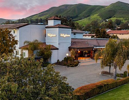A scenic hotel sits at the foot of green hills, with a light-blue facade, palm trees, and a welcoming driveway at sunset.
