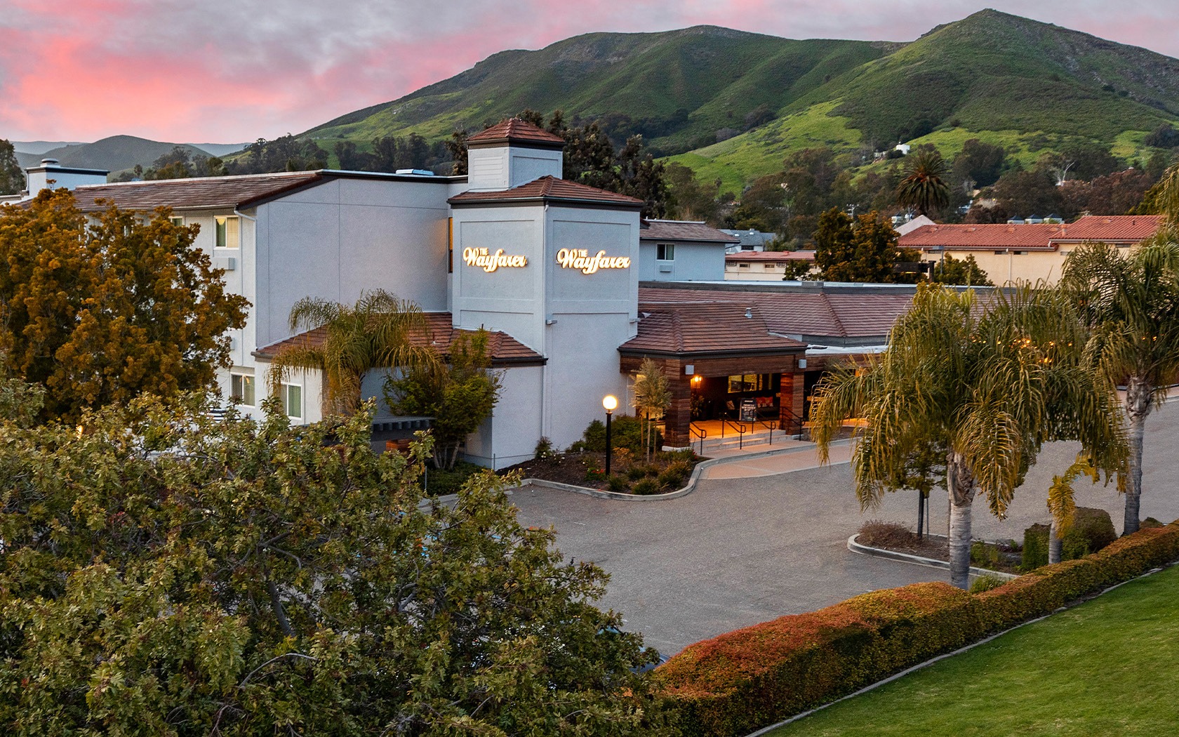 A scenic hotel sits at the foot of green hills, with a light-blue facade, palm trees, and a welcoming driveway at sunset.