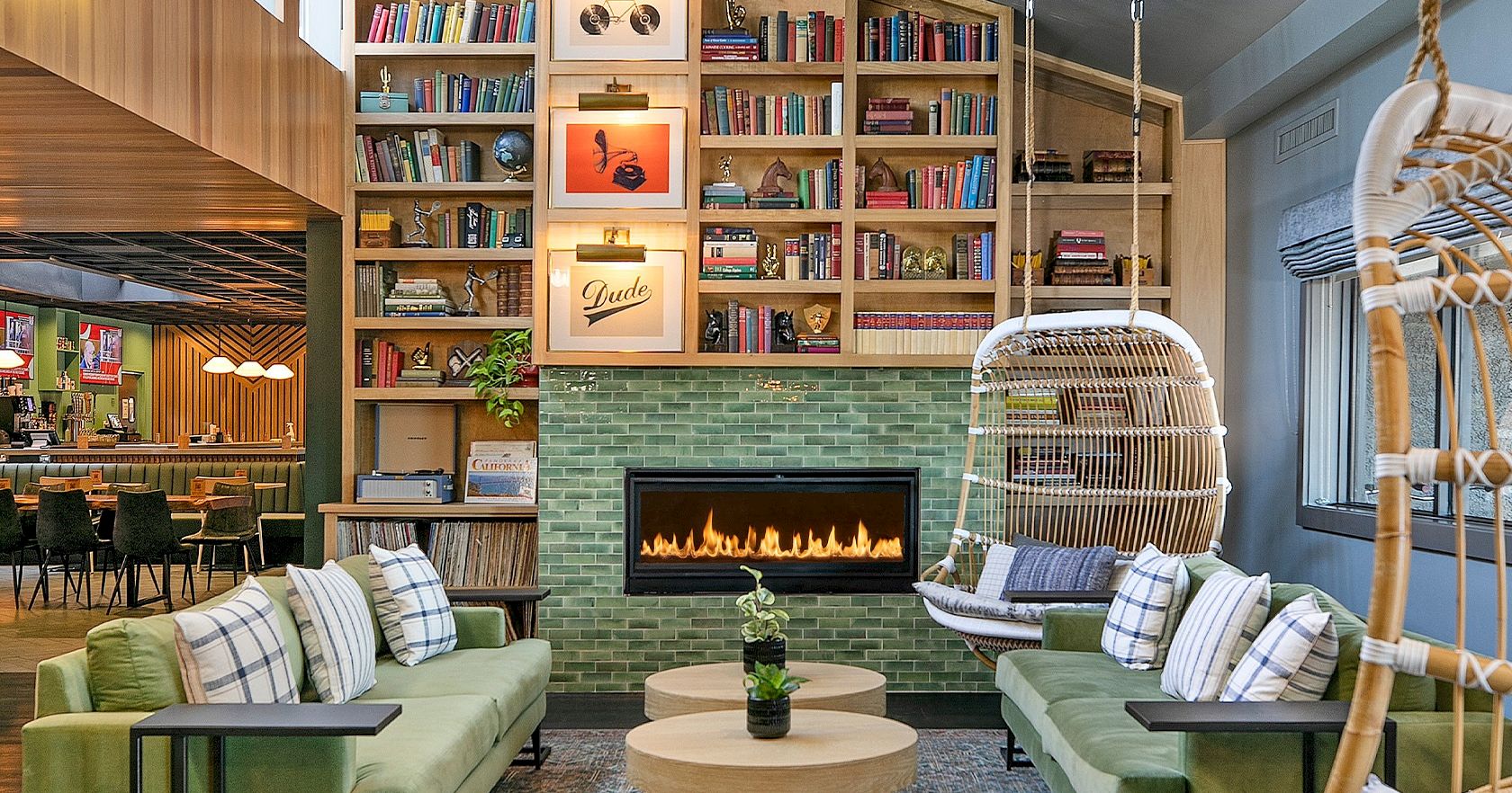 A cozy, modern library-lounge with green sofas, a brick fireplace, hanging chair, and tall book shelves filled with books, creating a warm communal vibe.