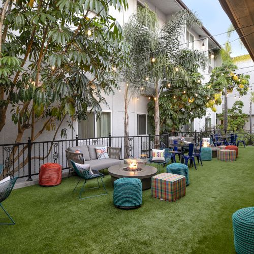 A cozy outdoor lounge with string lights, colorful poufs, low tables, and scattered seating on a grassy courtyard between buildings.