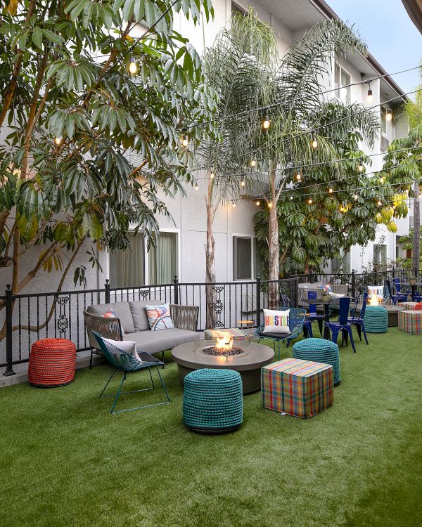 A cozy outdoor lounge with string lights, colorful poufs, low tables, and scattered seating on a grassy courtyard between buildings.