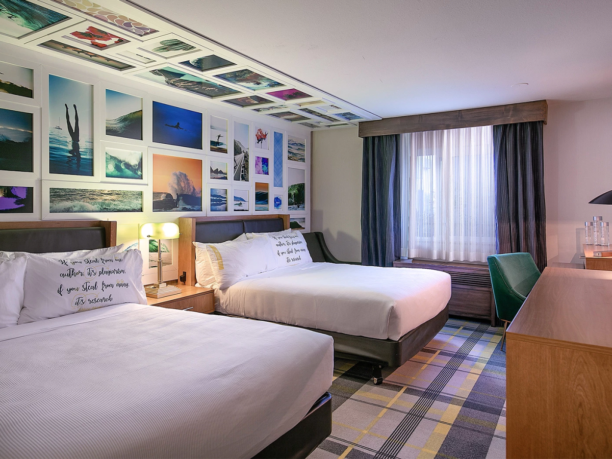 A hotel room with two queen beds, a wall collage of travel photos above, nightstands with lamps, a desk, a window with blue curtains, and a patterned carpet.