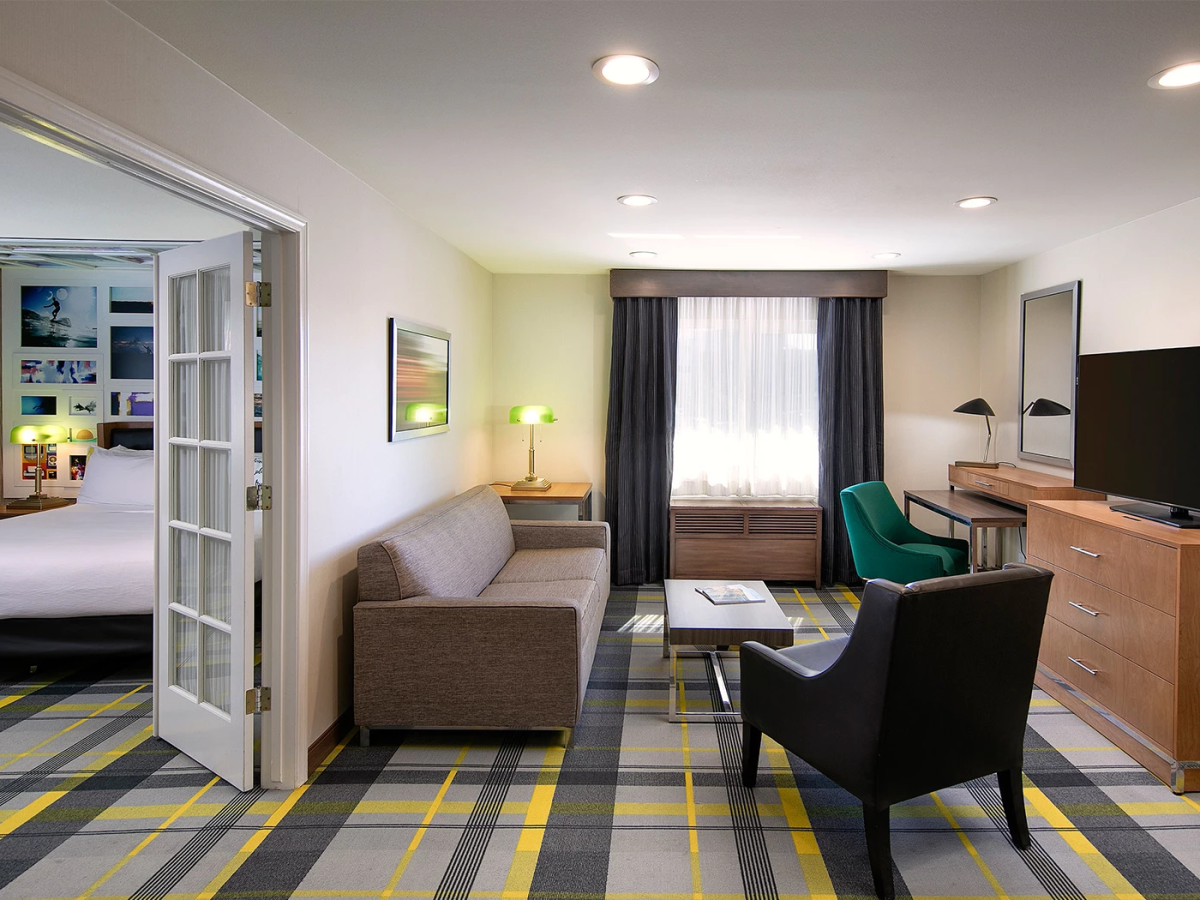 A hotel suite living area with a sofa, two chairs, coffee table, dresser with TV, and a doorway to a bedroom; window with dark curtains lights on.